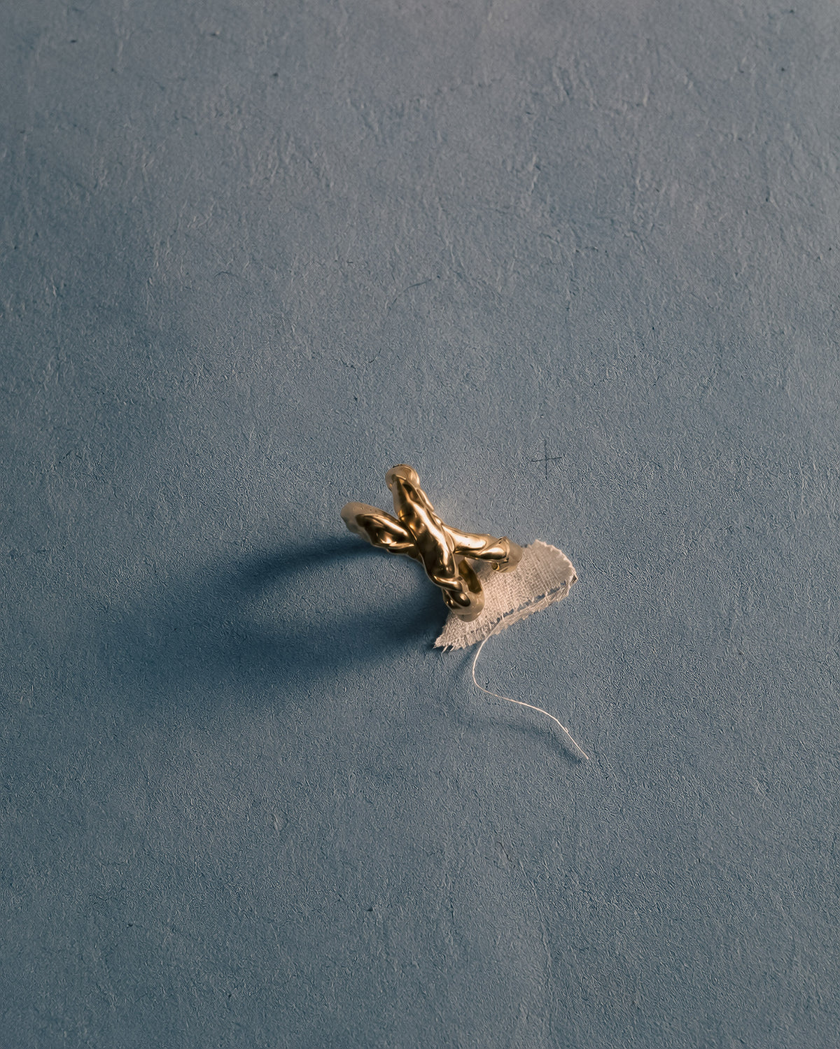 A recycled gold sculptural ring with a textured organic form and a torn white paper fragment, placed on a smooth slate-blue surface.