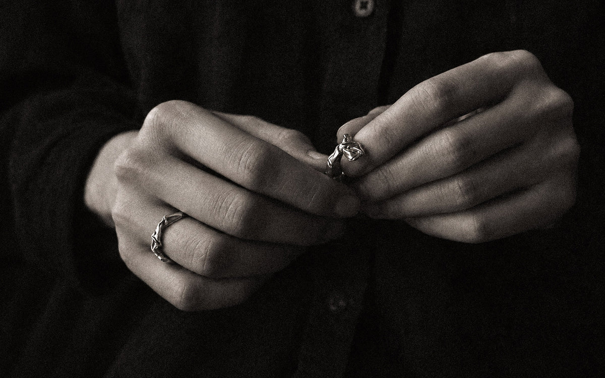 A close-up black and white photograph of a person's hands holding a silver ring, with another silver ring visible on the other finger. The person is wearing a dark button-up shirt.