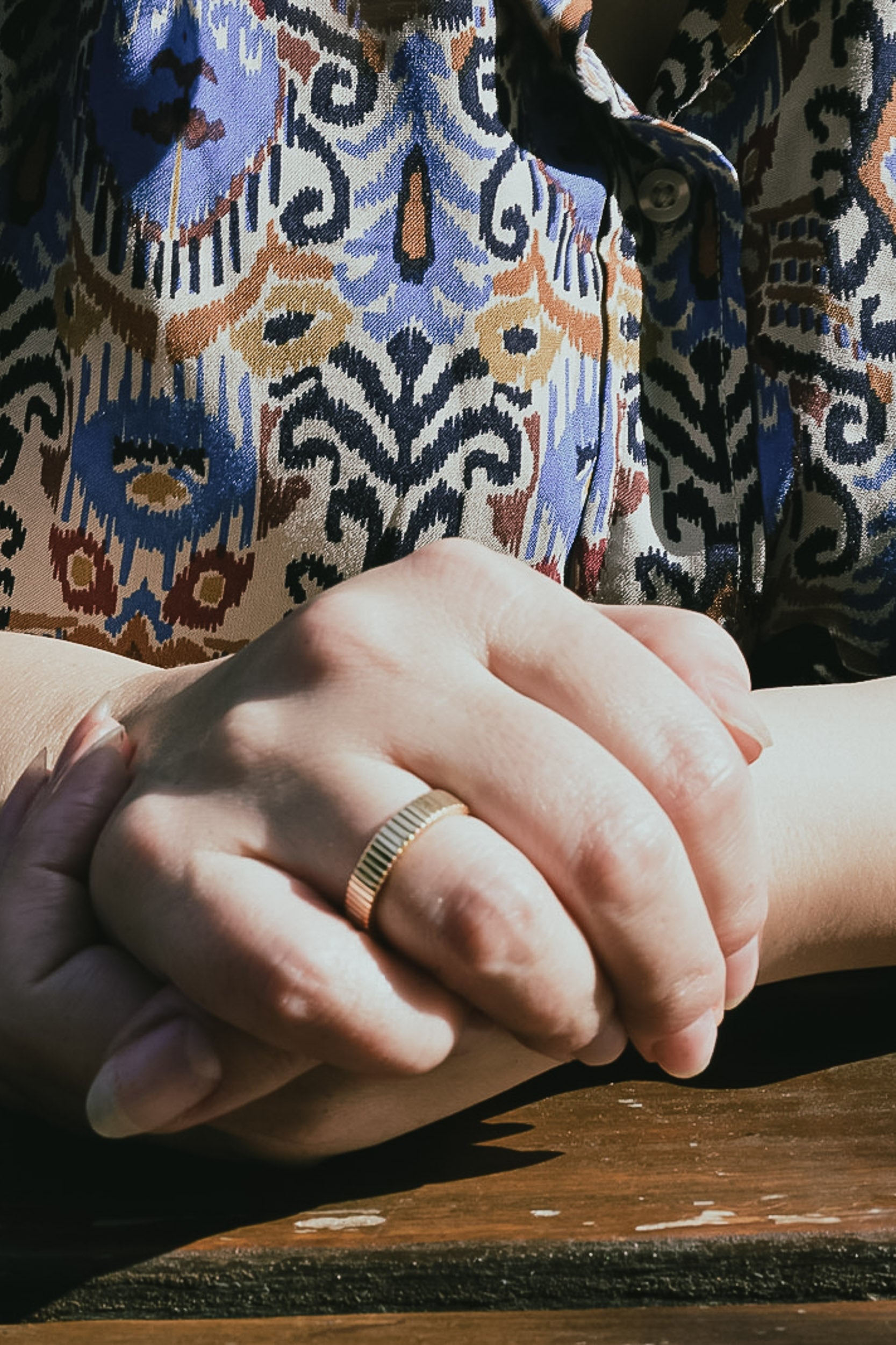 Close-up of two hands holding each other on a wooden surface, with one hand wearing a gold linear style ring.