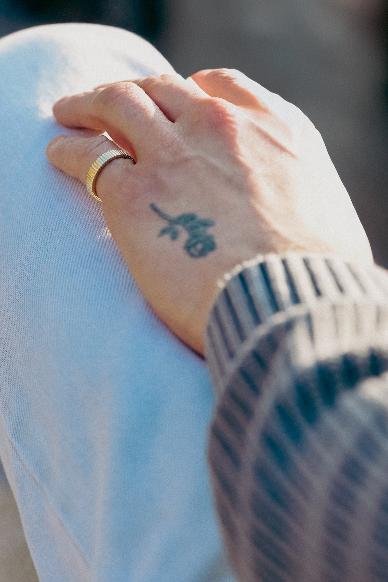 Hand with a tattoo on the wrist wearing a gold ring linear styled, against a blurred background.