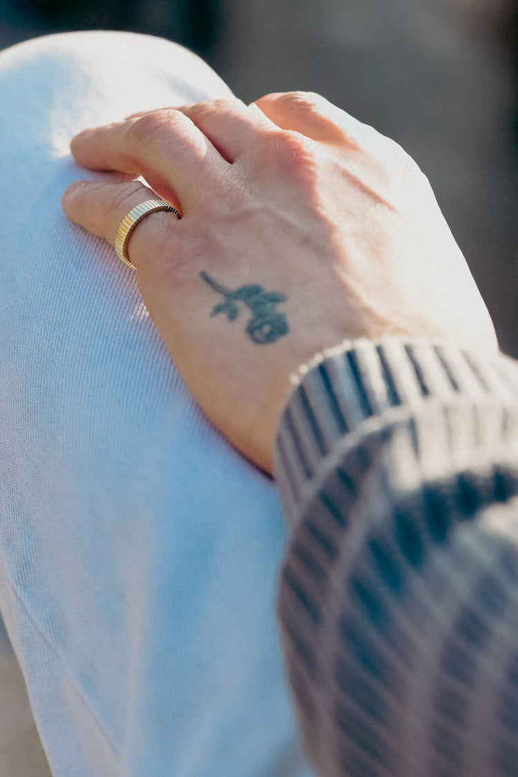 Hand with a tattoo on the wrist wearing a gold ring linear styled, against a blurred background.