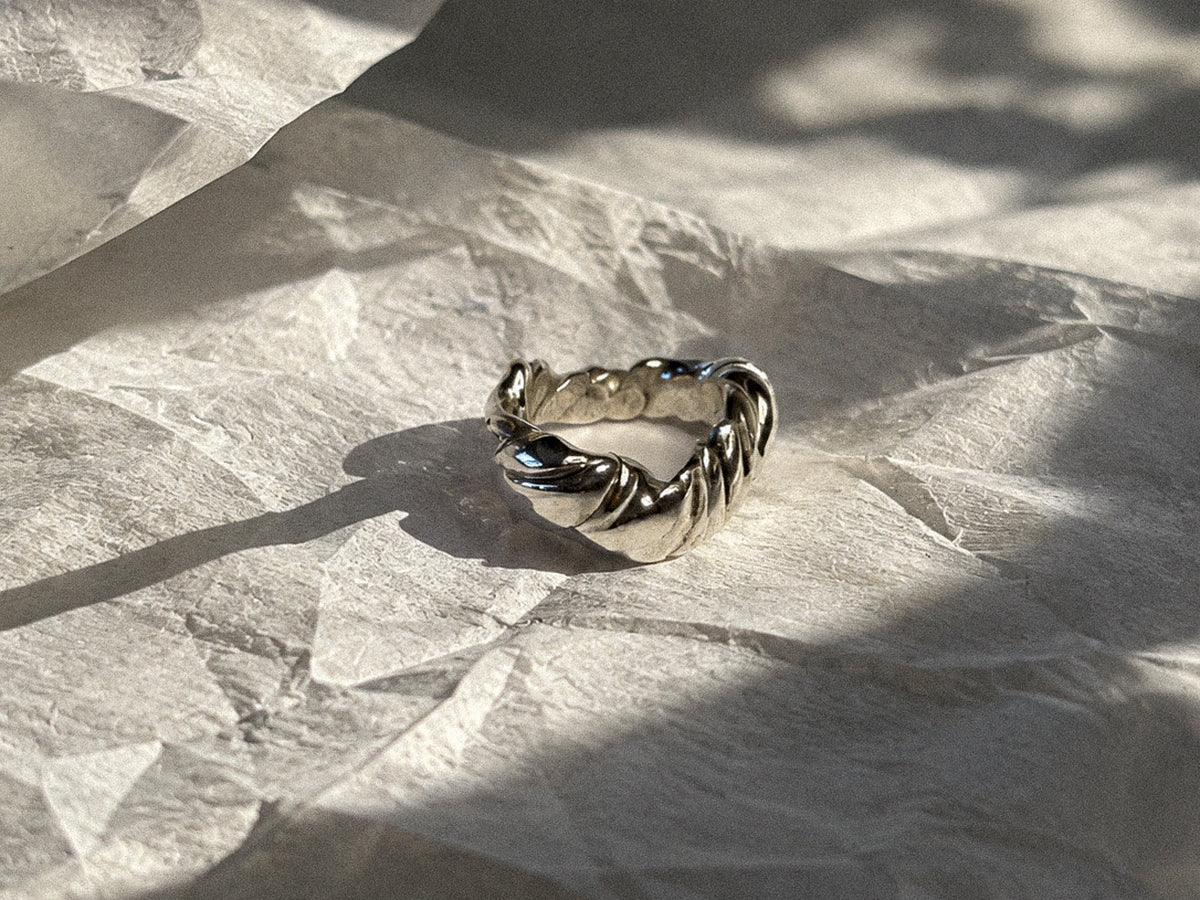 A twisted silver ring with a textured, organic like design, resting on crumpled white paper under dramatic, direct sunlight and shadows.