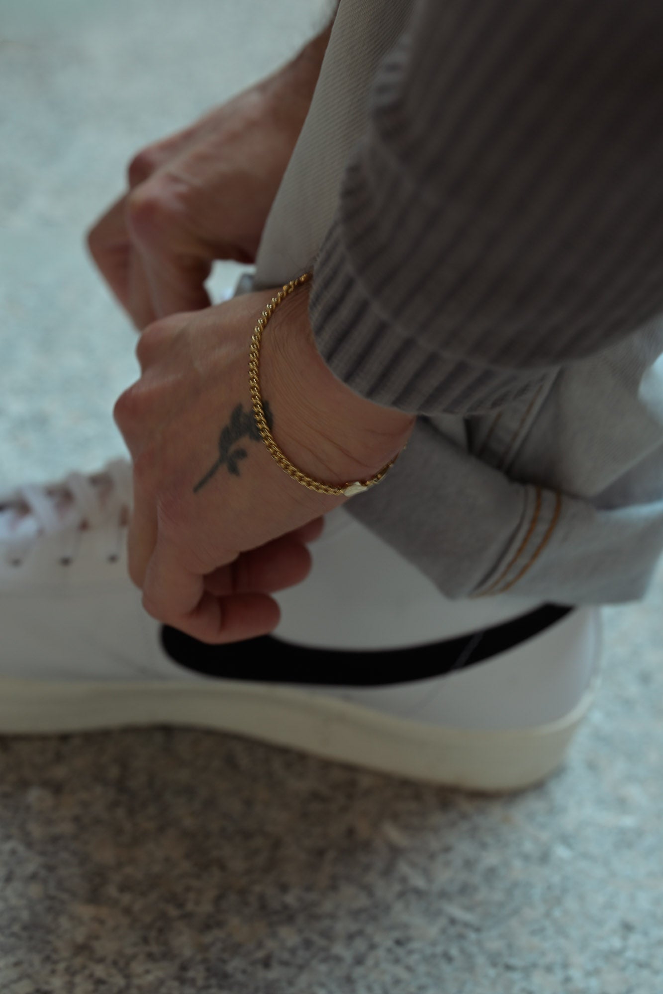 Close-up of a person's hands with a tattoo and gold curb style bracelet, wearing white sneakers aesthetic sneakers