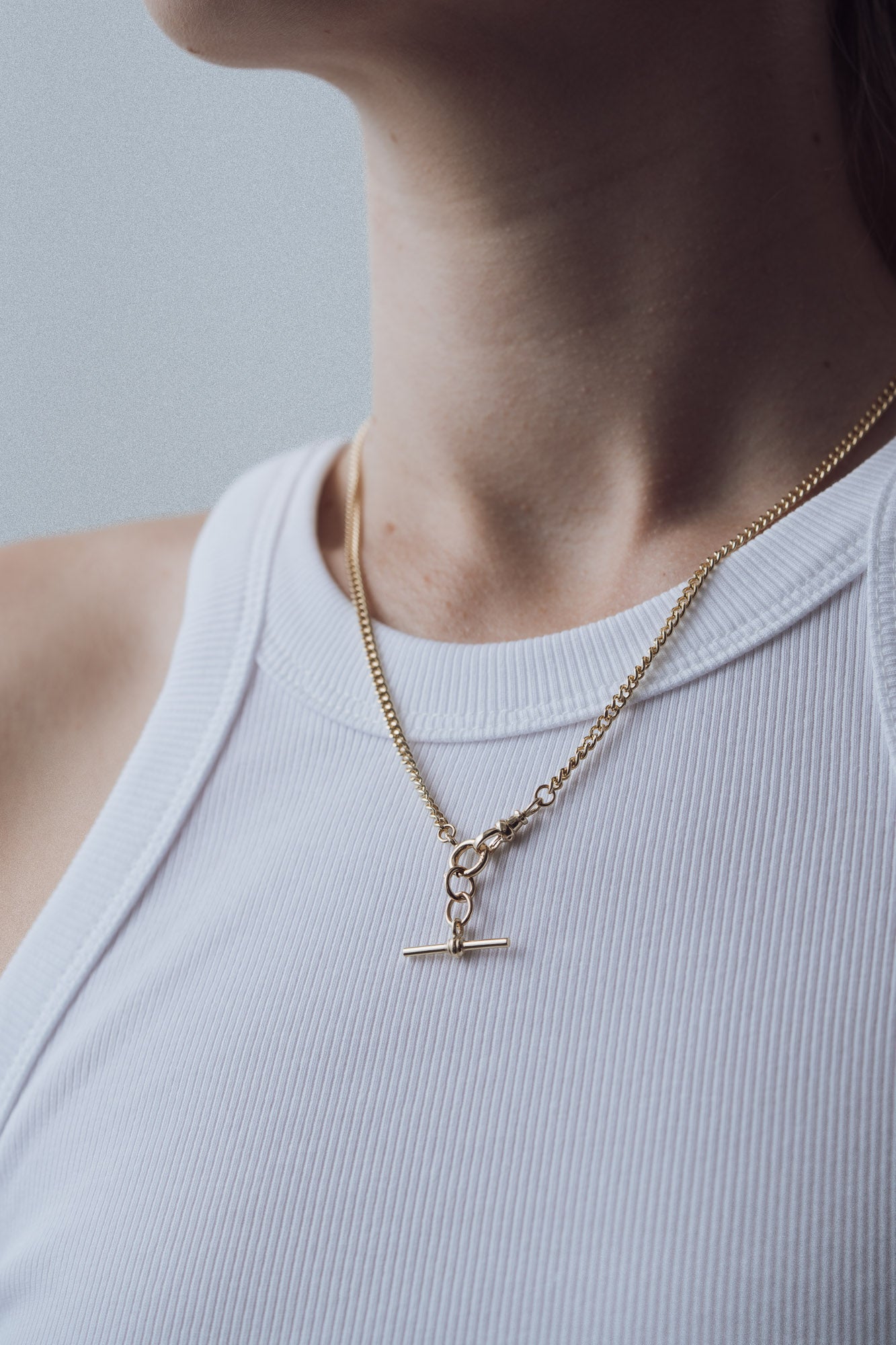 Gold necklace with a t bar pendant on a person wearing a light blue top against a neutral background