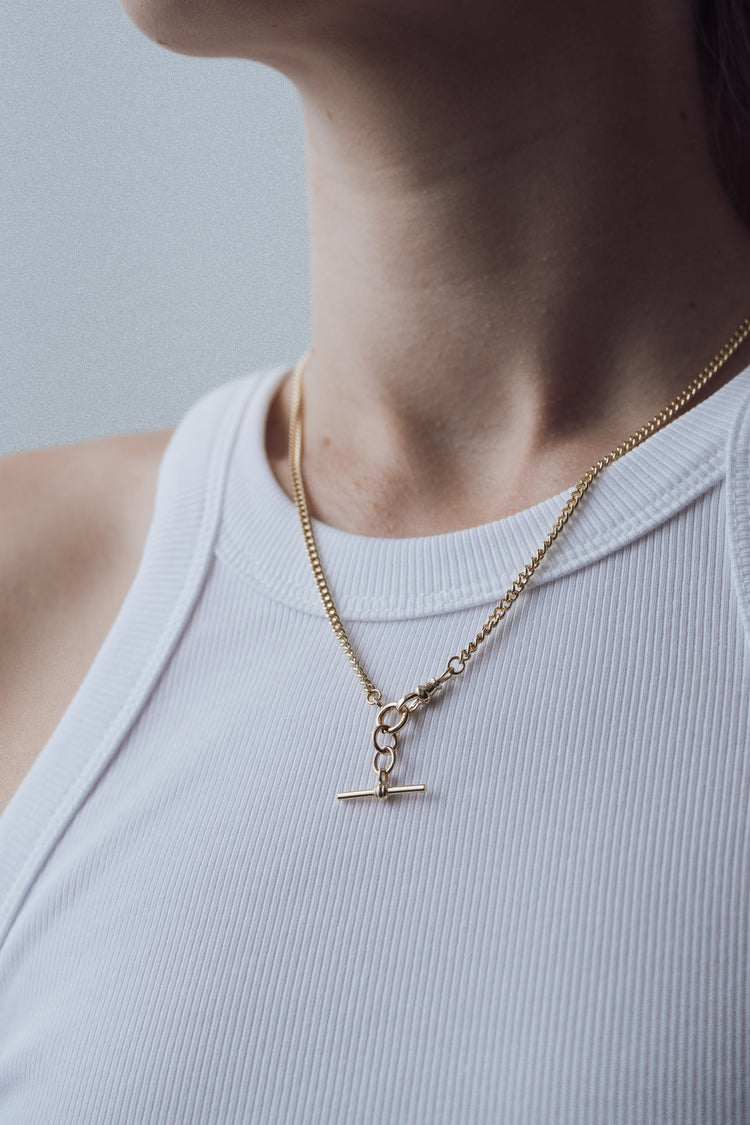 Gold necklace with a t bar pendant on a person wearing a light blue top against a neutral background