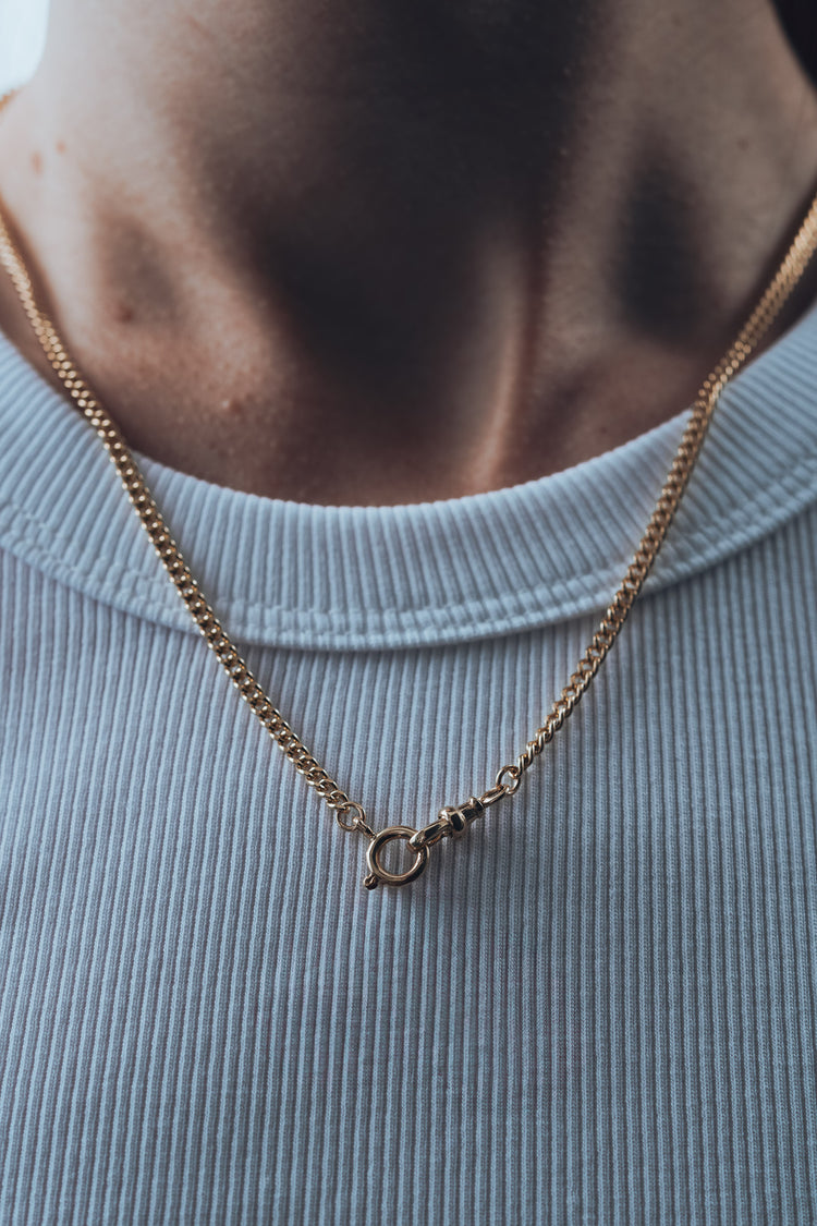 Gold chain necklace with a t bar pendant on a person wearing a light blue shirt
