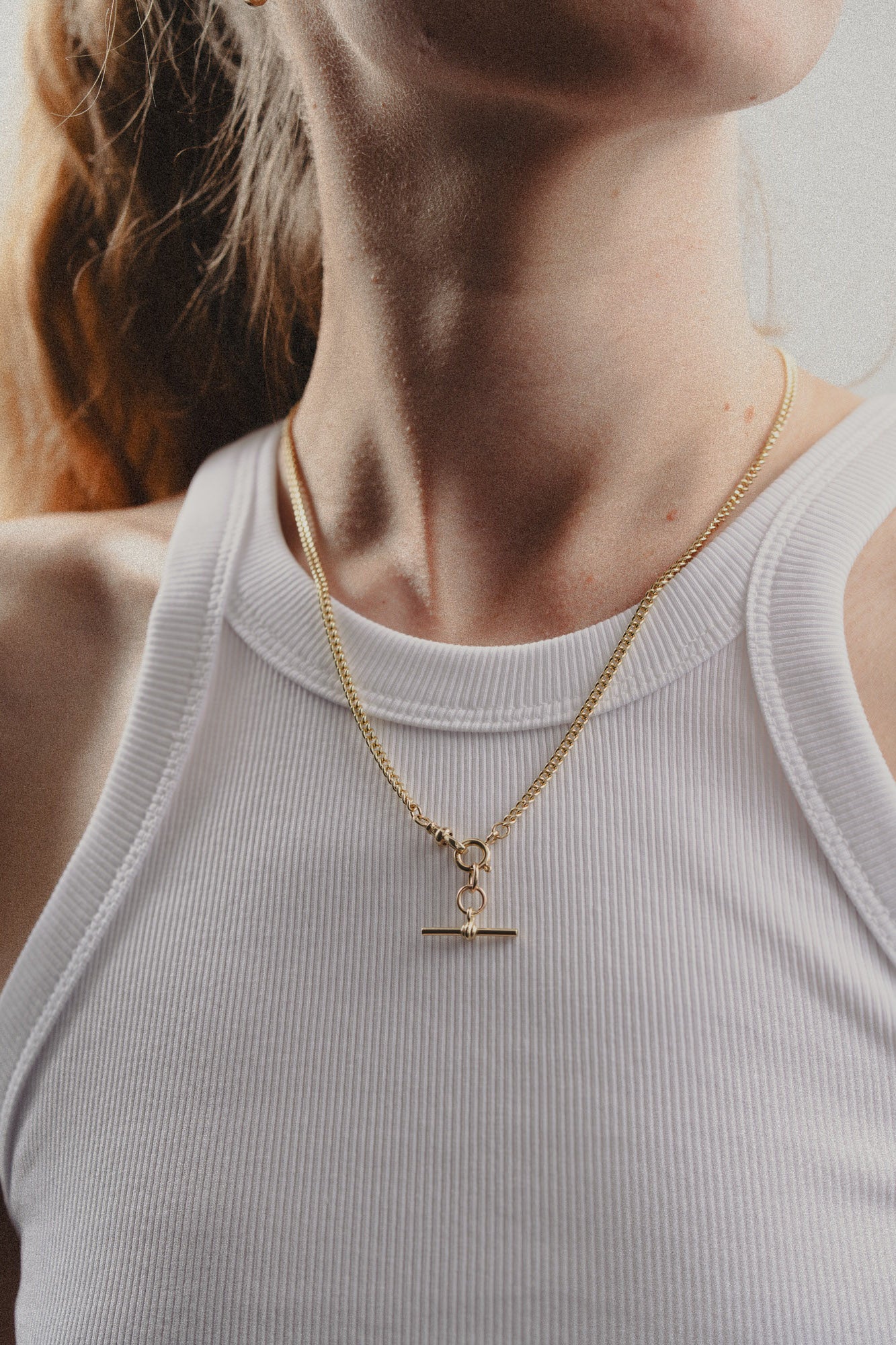 Gold necklace with a T bar pendant worn by a person in a light gray tank top.